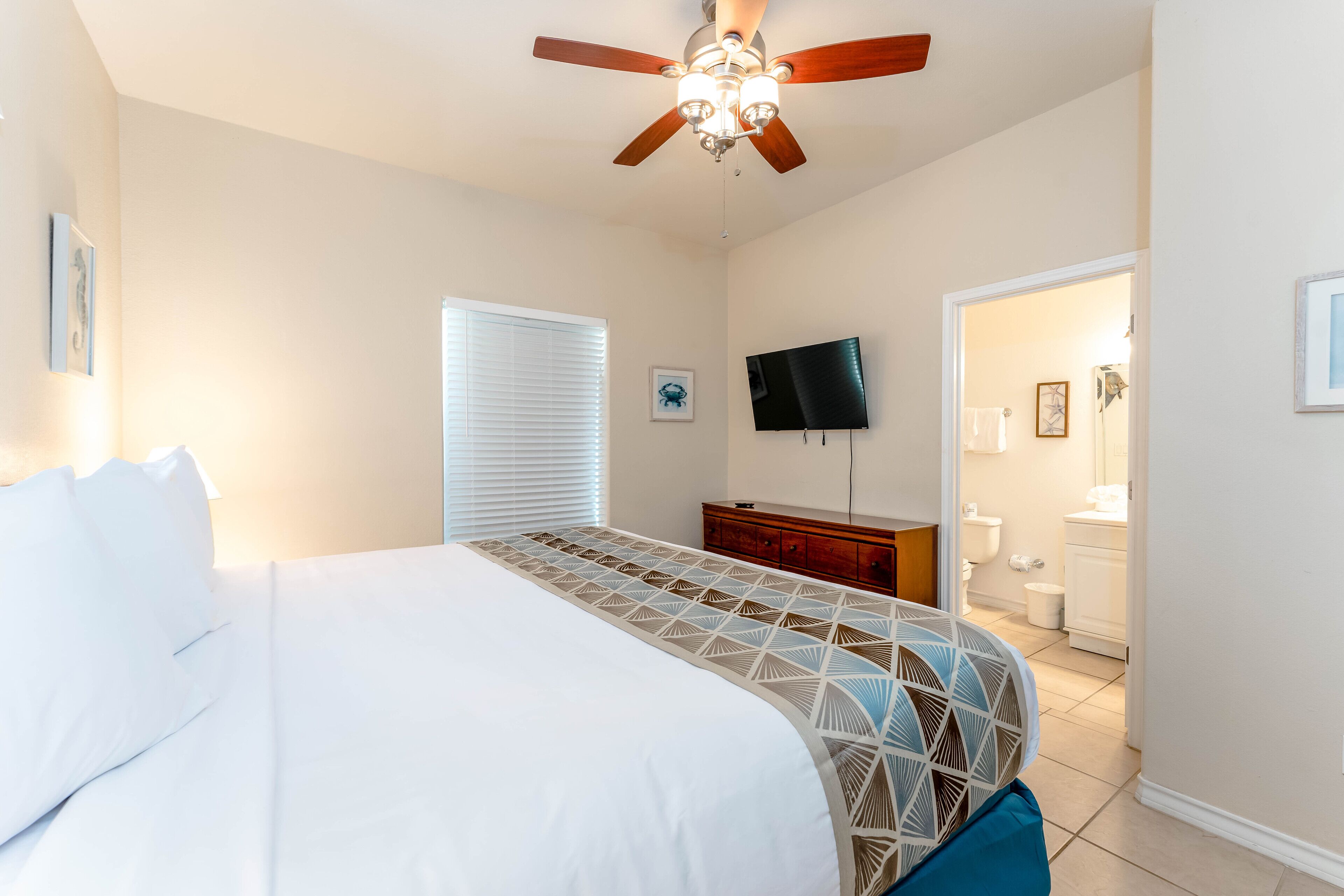 Une chambre à coucher avec un lit, un ventilateur de plafond, une télévision et une salle de bain visible à travers une porte ouverte.