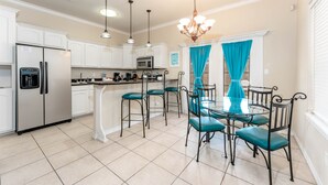 Una cocina moderna con armarios blancos, electrodomésticos de acero inoxidable, sillas color turquesa y una mesa de comedor de vidrio.