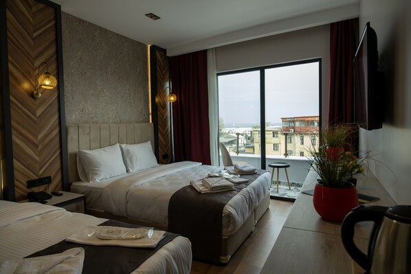 A hotel room with a large bed, a smaller bed, a desk with a telephone, a red potted plant, and a view of buildings through the window. - Linda Port Hotel (Istanbul)
