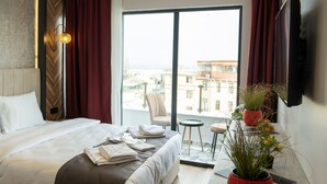 Quarto de hotel moderno com uma cama grande, uma mesa pequena, uma planta em vaso e vista para a cidade.