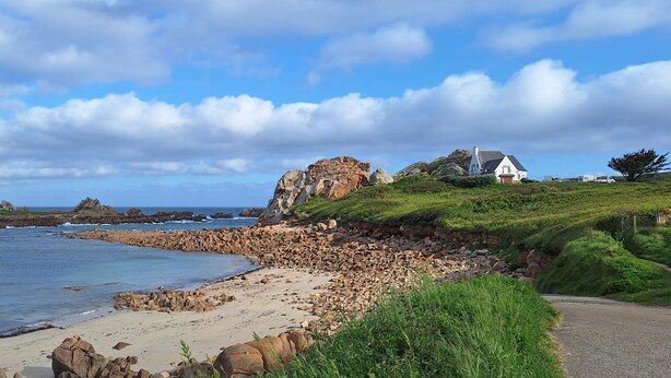 A coastal landscape with a rocky beach, a white house with a black roof, and a clear sky.