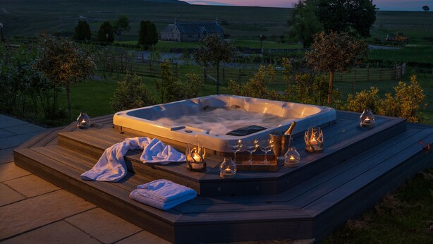 Un jacuzzi con escalones, rodeado de velas y vasos, sobre una terraza de madera con vistas panorámicas al campo.