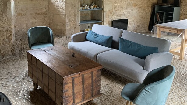 A rustic room with exposed wooden beams, a stone wall, a sofa with cushions, a wooden coffee table, a chair, and a shelf with items. - Charmante Maison Périgourdine (Granges-d'Ans)