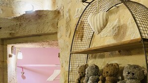 Una habitación con paredes de piedra, una puerta rosa, una estantería metálica con osos de peluche y una lámpara blanca.