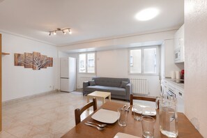 A modern living room with a grey sofa, a dining table set with plates, glasses, and cutlery, and a kitchen area with a microwave and a washing machine.