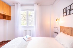 A hotel room with a bed, a radiator, a window with curtains, a wooden headboard, and a wall-mounted light fixture.
