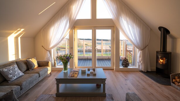 A spacious living room with a high ceiling, a large window with curtains, a sofa, a coffee table, and a wood-burning stove. - Luxury two bedroom converted steading (Perth)