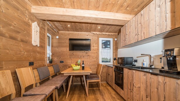 A cozy cabin-style room with wooden walls and floors, a dining area with benches, a kitchen with modern appliances, and a TV mounted on the wall. - 1214 Piz Buin Schima Drosa (Gaschurn)