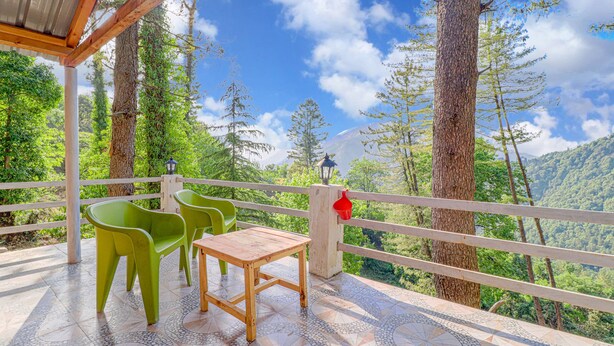 A terrace with a wooden table and green chairs, overlooking a forested mountain area.