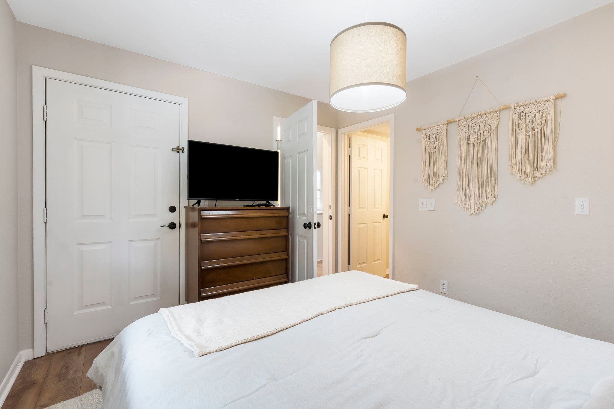 Une chambre à coucher comprenant un lit, une commode, un téléviseur et des décorations murales en macramé.