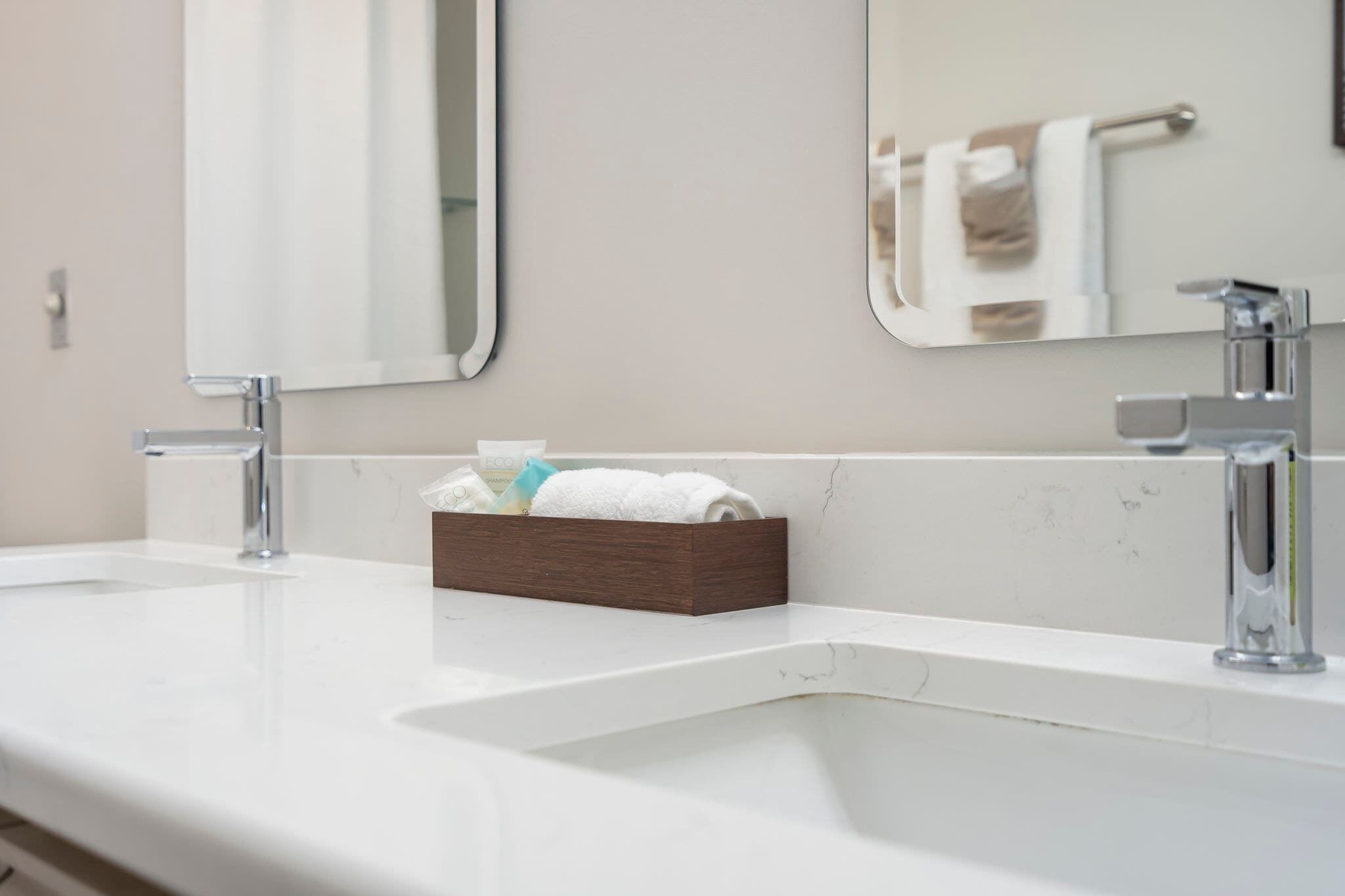 A bathroom with a white sink, two chrome faucets, a wooden box with toiletries, and a mirror reflecting a towel rack.