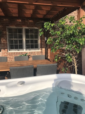 A hot tub with a wooden deck and gray chairs, a brick wall, and a window with blinds.