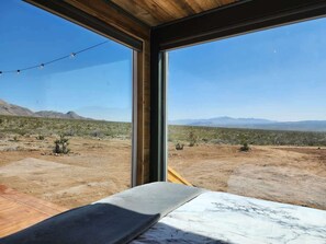 Ein Zimmer mit großem Fenster und Blick auf eine Wüstenlandschaft, in der Ferne Berge.