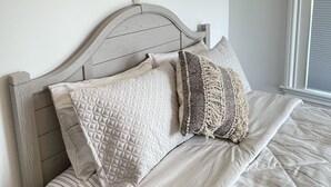 Un lit bien fait, avec une couvre-lit à rayures et plusieurs coussins, dont une décoration murale en macramé au-dessus.