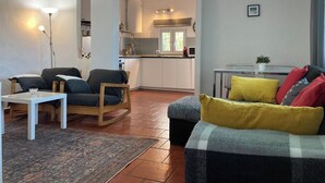 A living room with a sofa, chairs, a small table, and a rug. There is a kitchen in the background with cabinets and a window. - T1 Sunset View (Albufeira)
