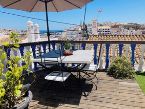 A balcony with a table and chairs, a potted plant, and a view of the cityscape and sea. - T1 Sunset View (Albufeira)