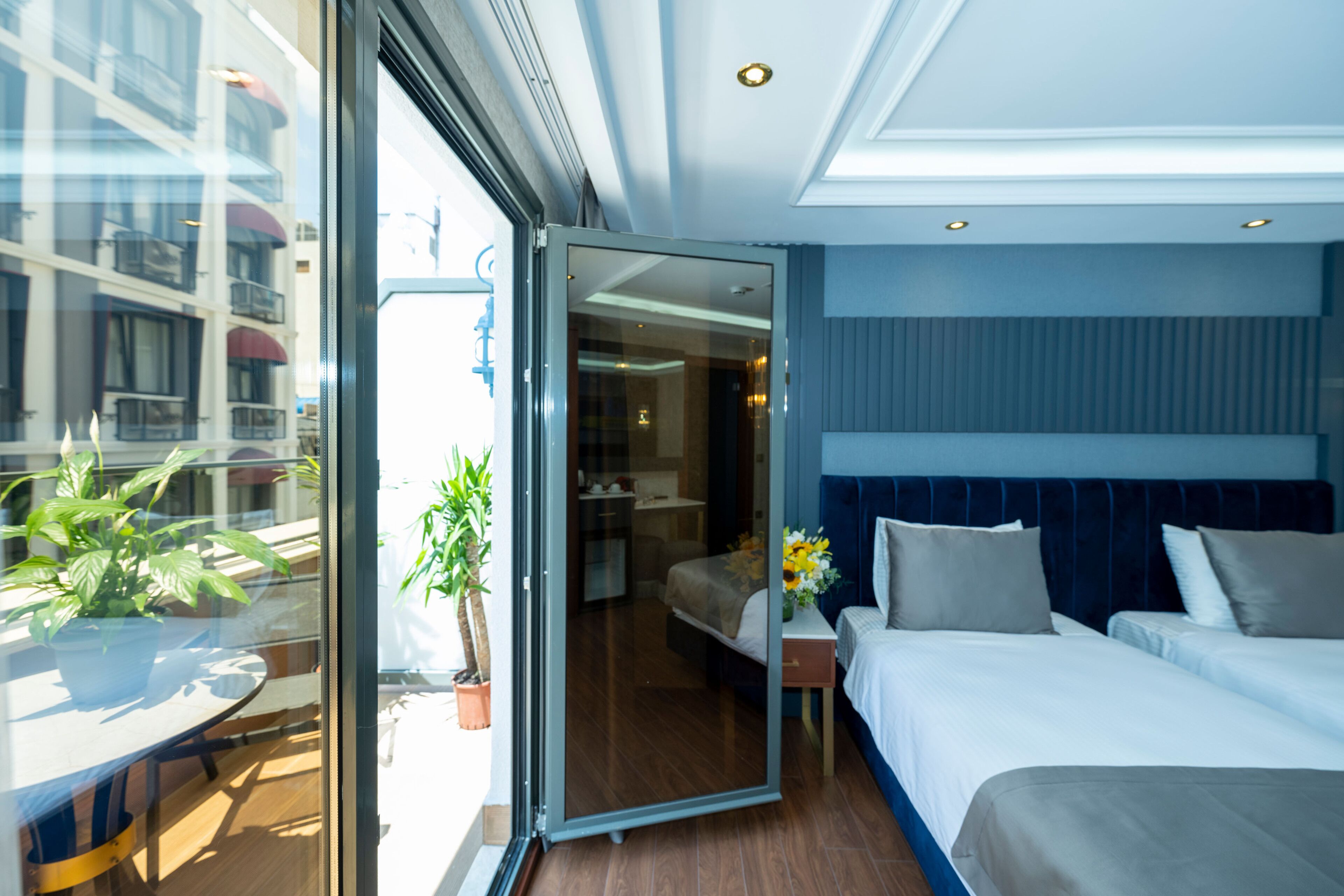 A modern hotel room with a large bed, a small table with a potted plant, and a glass door leading to a balcony with a view of a building.