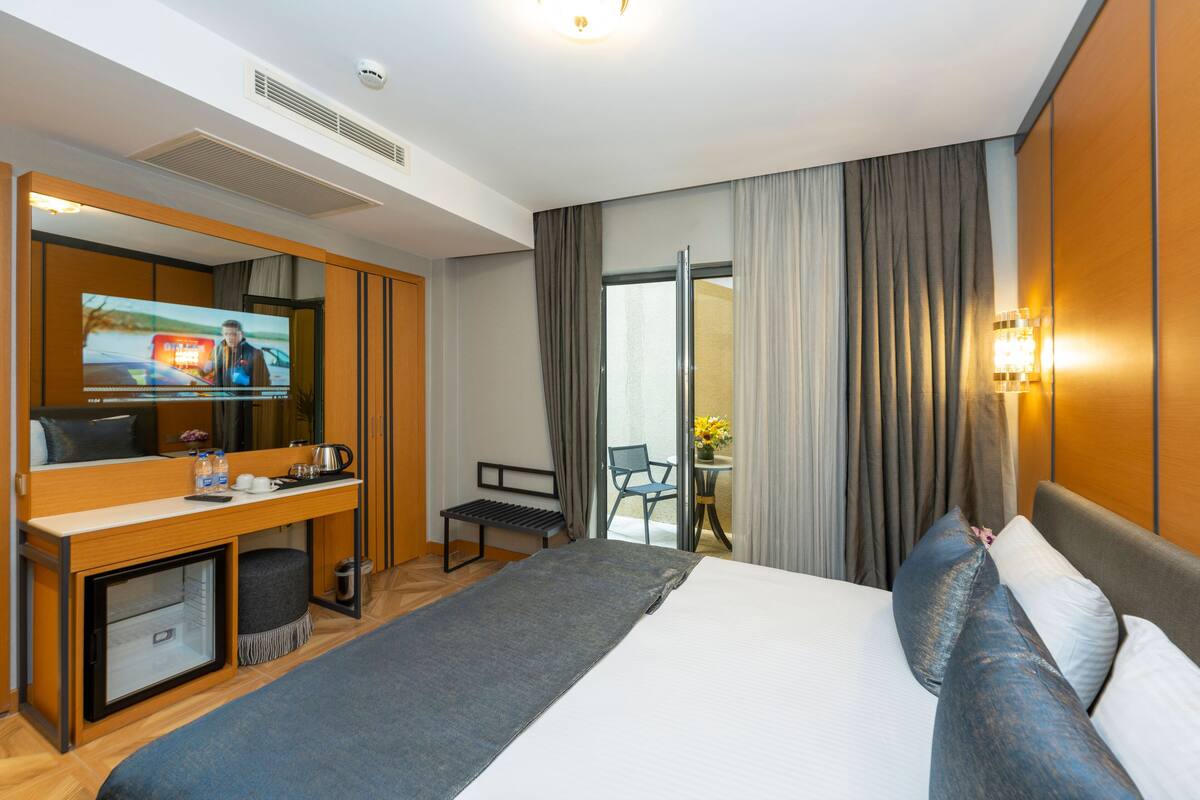 A hotel room with a bed, a desk with a television, a small table with a chair, and a sliding glass door to a balcony.
