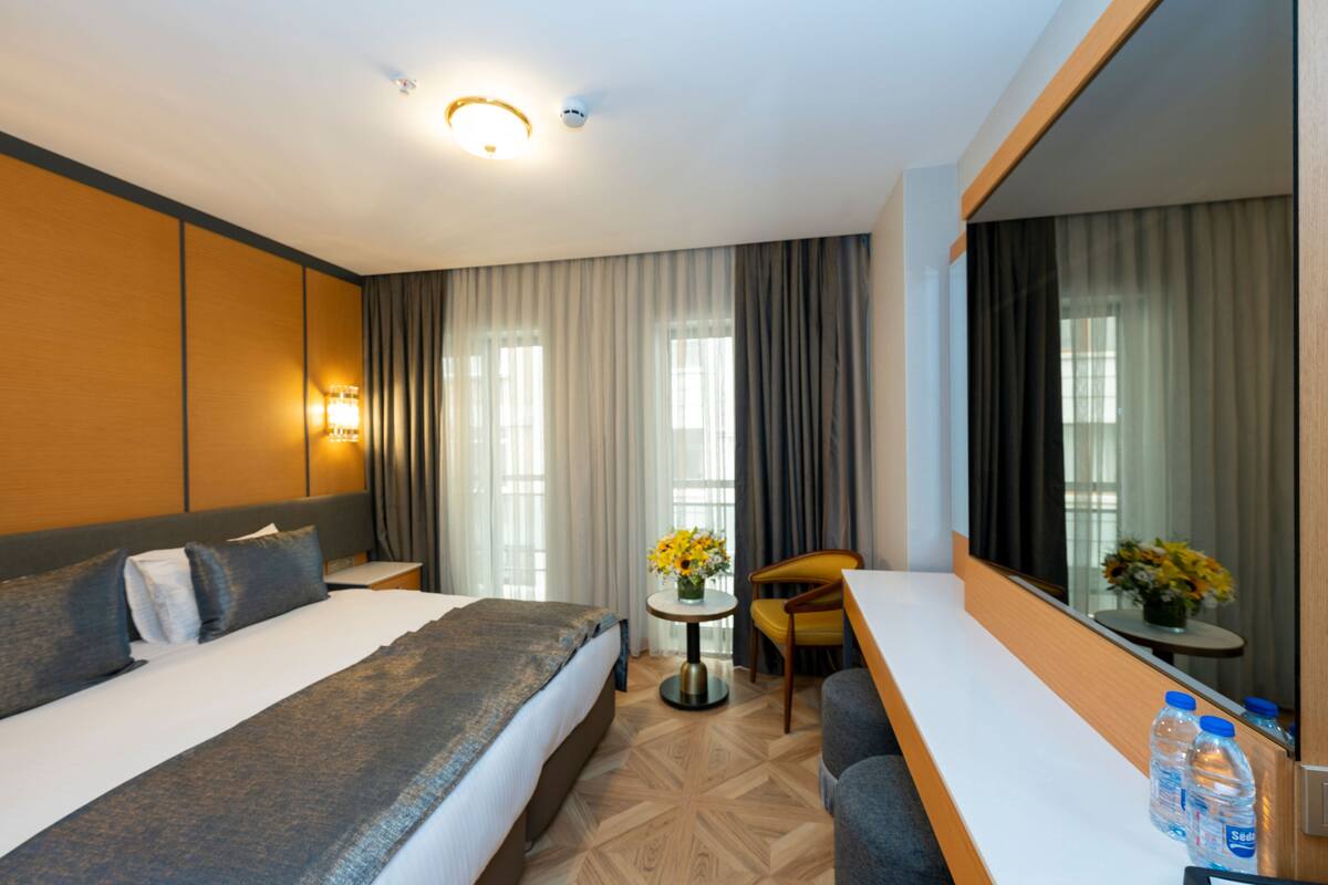 A hotel room with a large bed, a desk, a chair, a small table with flowers, and a mirror.