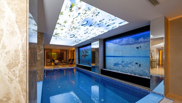 Una piscina interior moderna con techo de cristal, pantallas grandes que muestran escenas submarinas y un área de descanso con sillones reclinables.