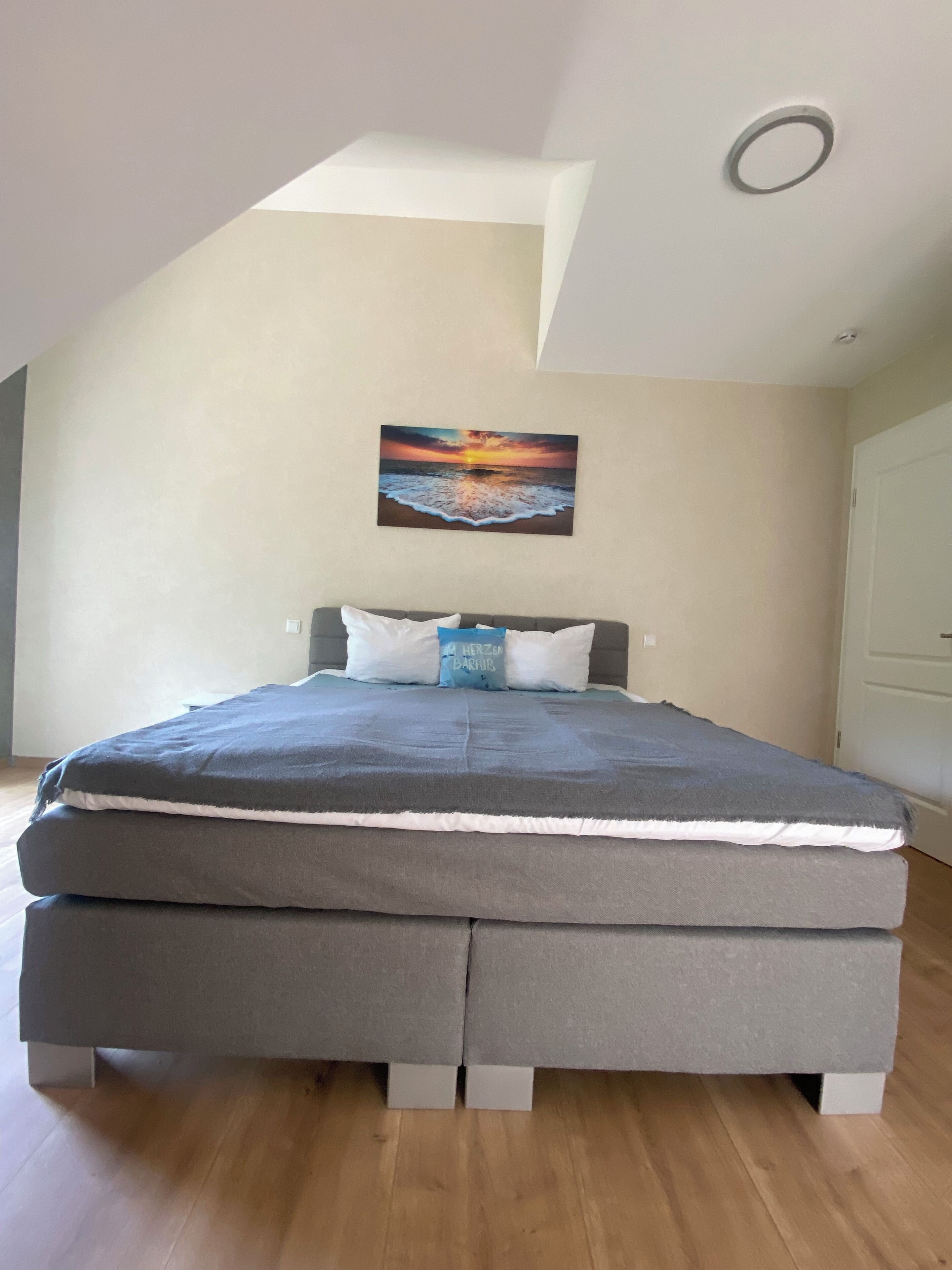 A bedroom with a bed, pillows, and a painting of a sunset over the ocean.