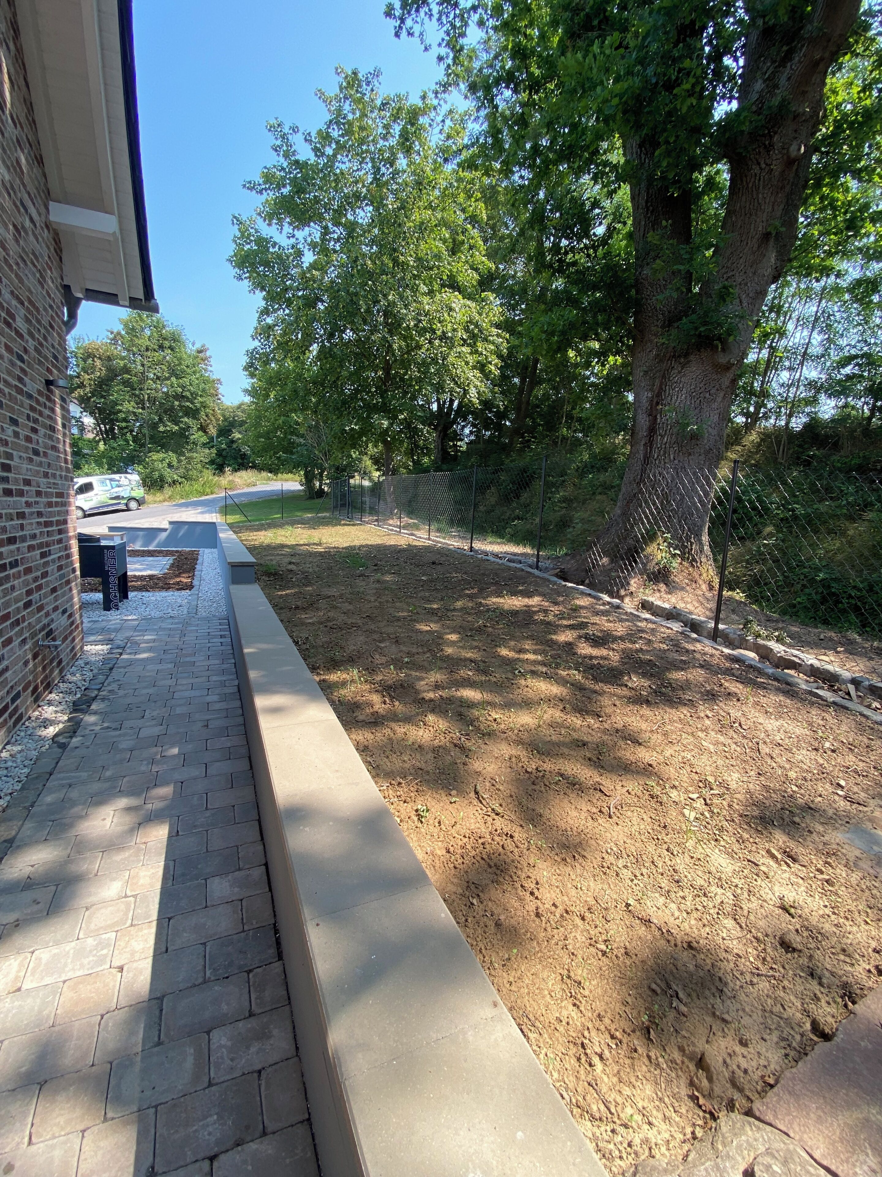 A paved pathway adjacent to a brick building, a fenced area with trees, and a dirt patch.