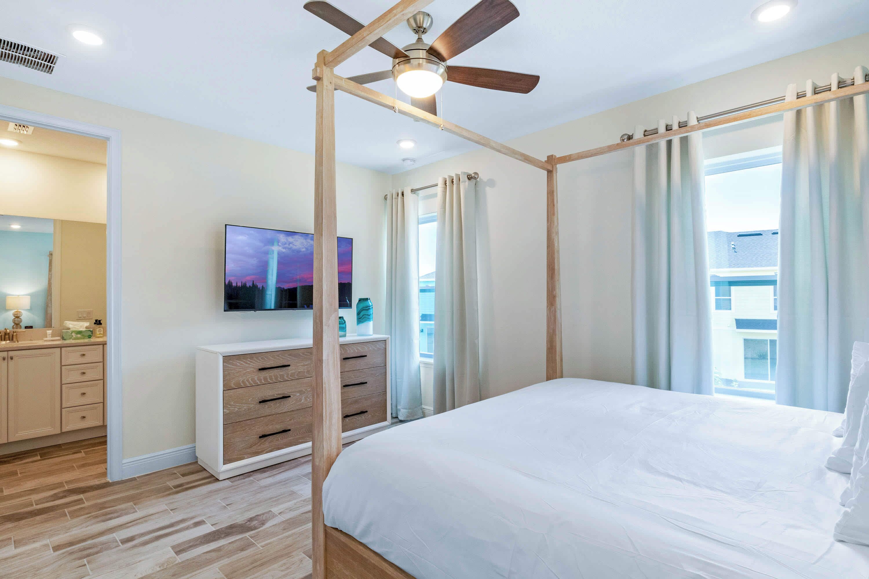 Un dormitorio con cama, ventilador de techo, televisor, cómoda de madera y ventana con cortinas.