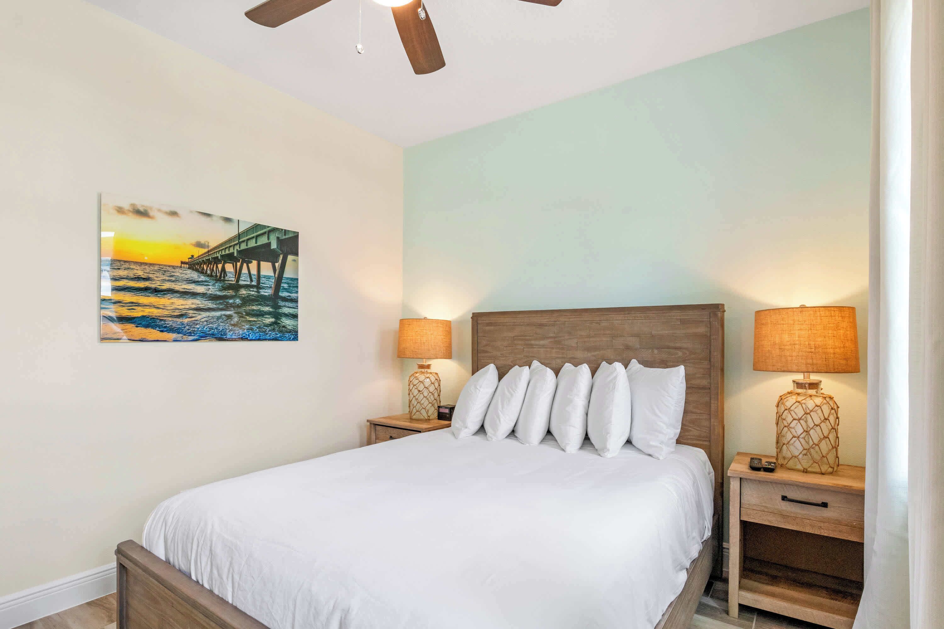Un dormitorio con cama, dos mesitas de noche, un ventilador de techo, un cuadro de un muelle y una pared de color claro.
