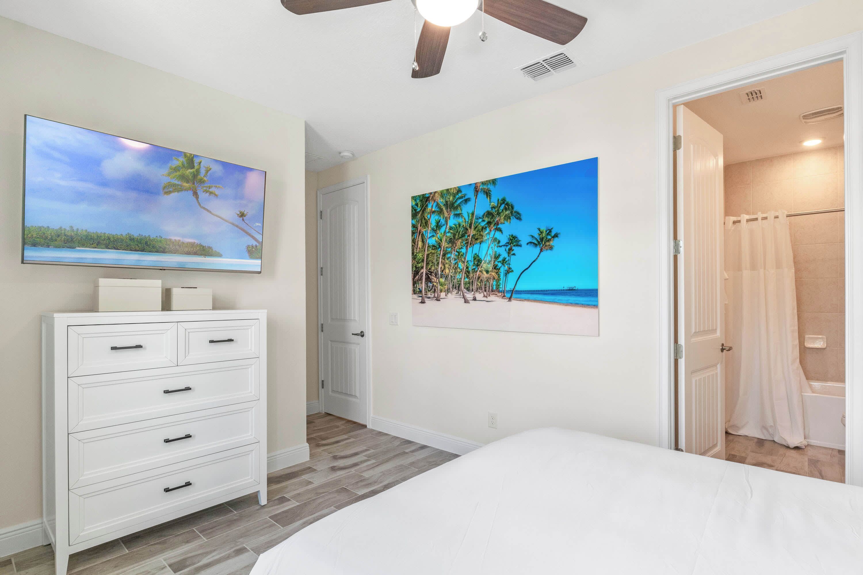 Een slaapkamer met een bed, een televisie, een dressoir, een deur naar een badkamer en twee decoratieve panelen met tropische taferelen.