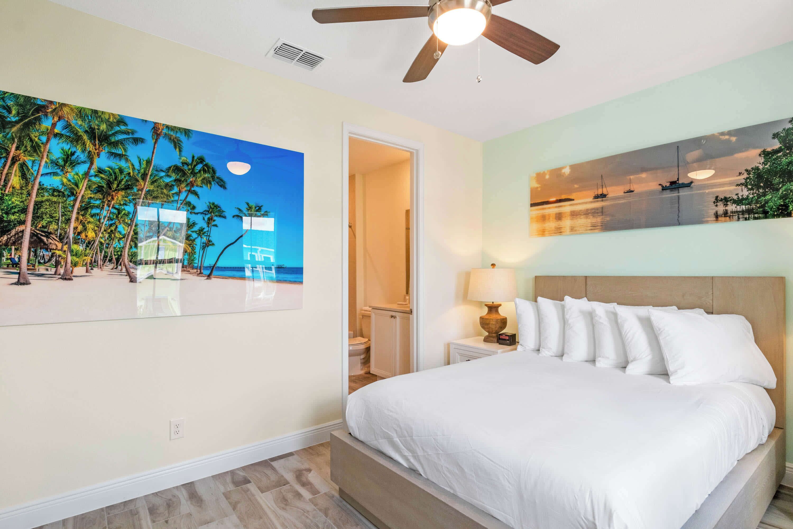 A bedroom with a bed, a ceiling fan, a nightstand with a lamp, and a painting of a beach scene.