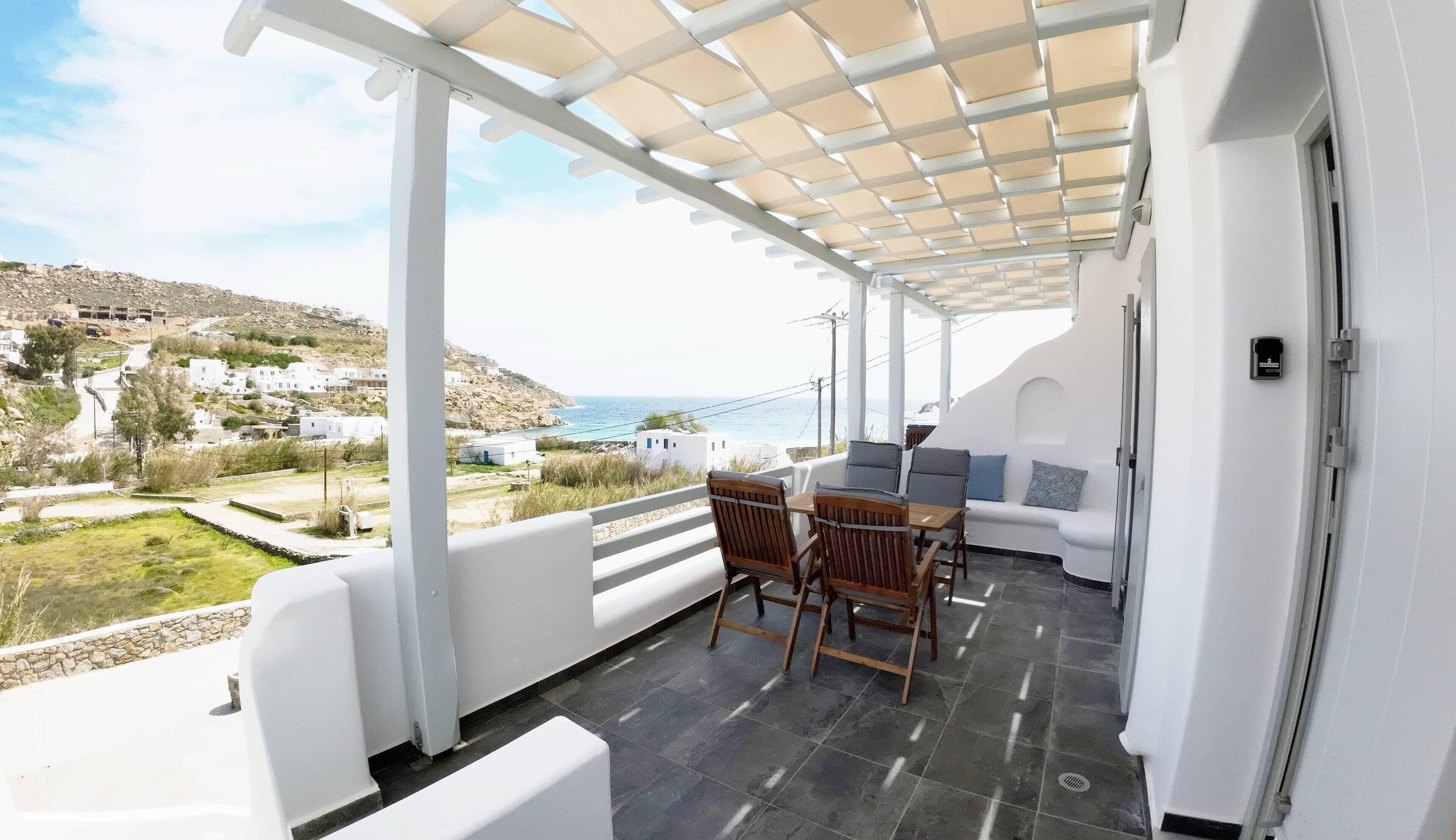 A covered outdoor area with a table and chairs, a sofa, and a view of the sea and buildings in the distance.