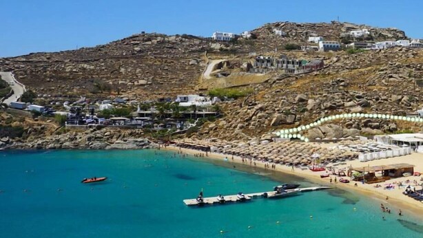 A coastal town with buildings on a hillside, a beach with umbrellas, and boats in the water. - Air house          . (Mykonos)
