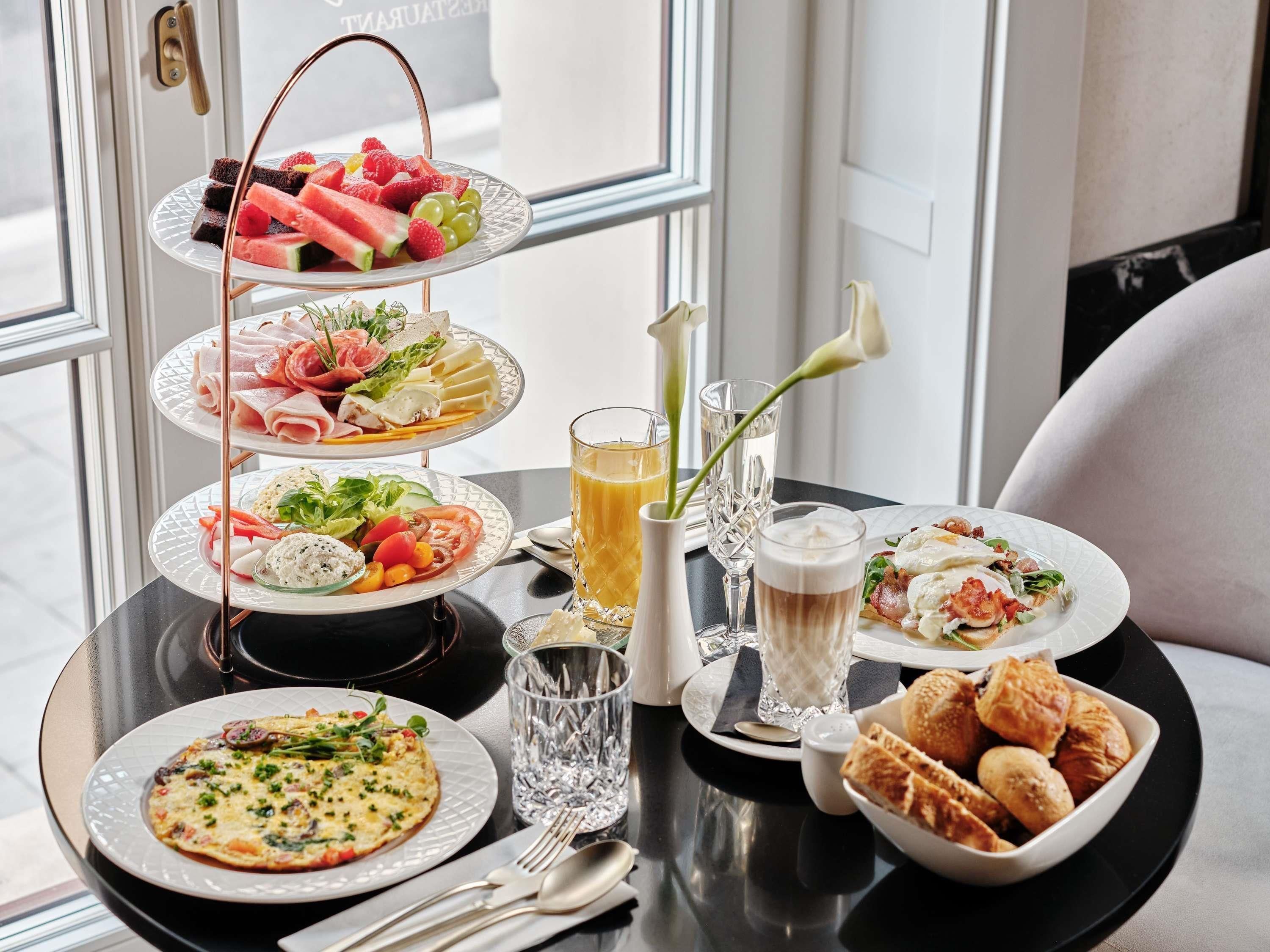 A breakfast buffet with various dishes, glasses of beverages, and a flower arrangement on a table by a window.