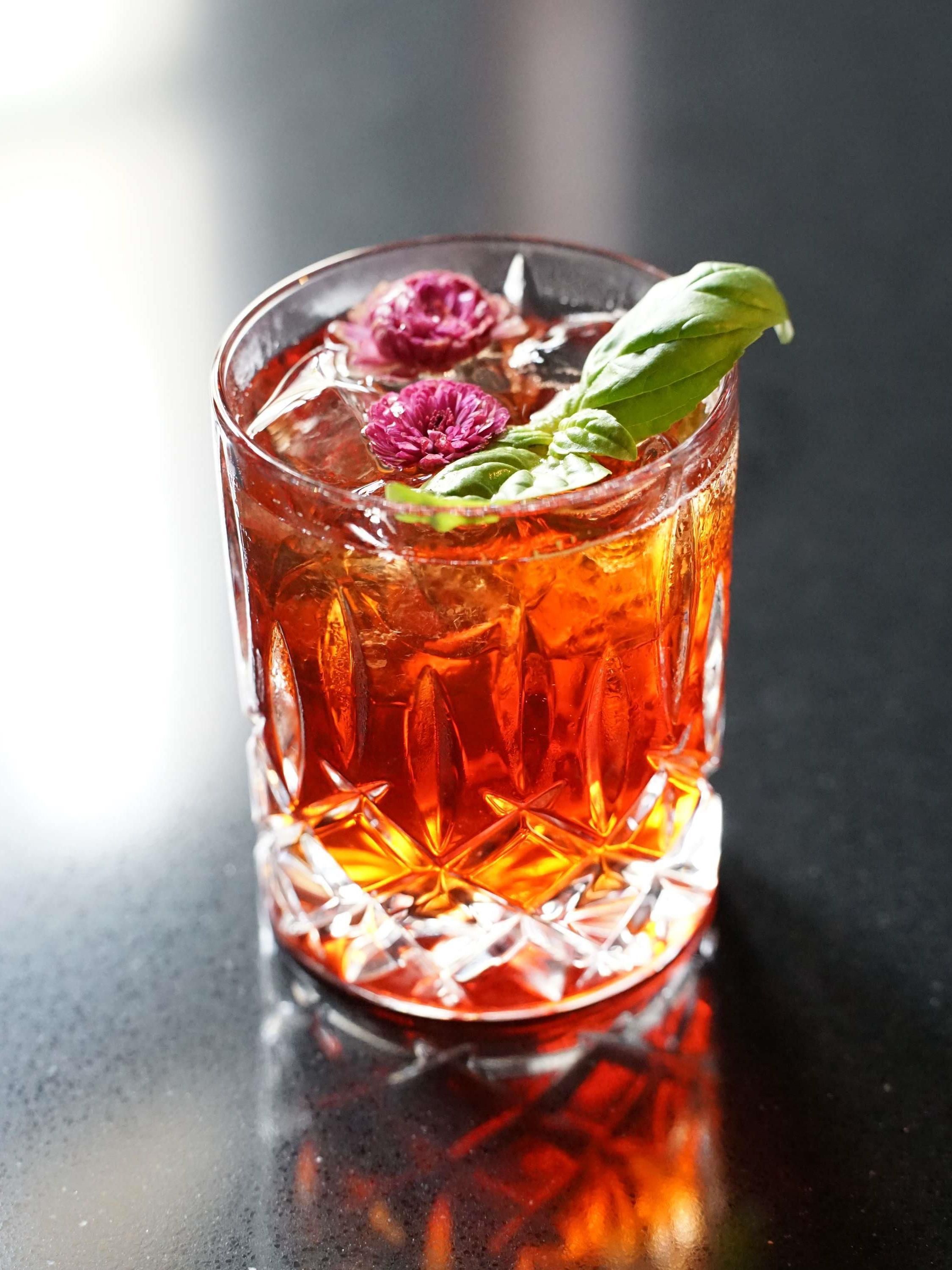 Un vaso transparente con un diseño texturizado, lleno de un líquido rojo, cubitos de hielo y adornado con una flor y una hoja.