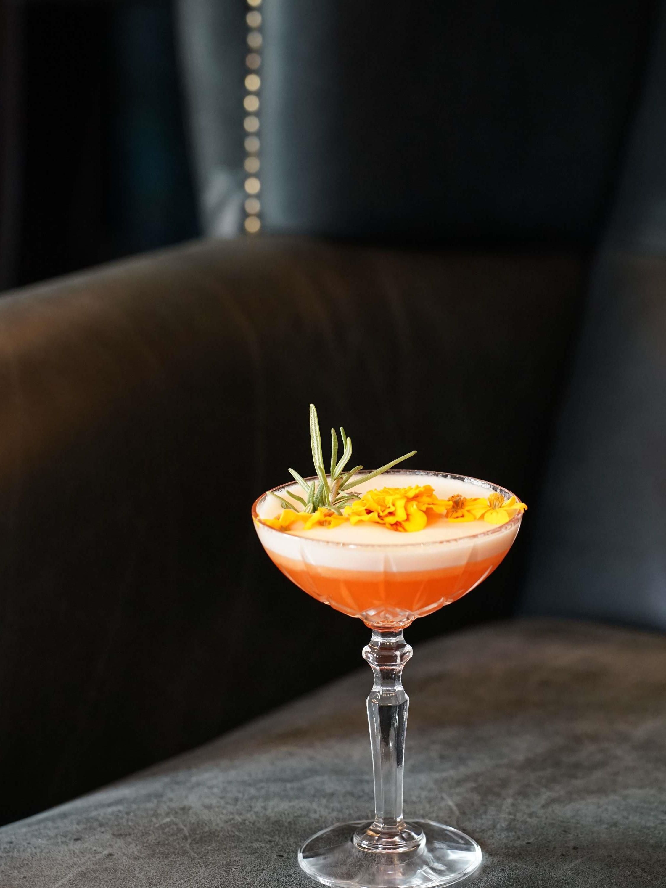 A cocktail in a stemmed glass with a layered drink, garnished with a sprig of rosemary and yellow flowers, on a dark surface.