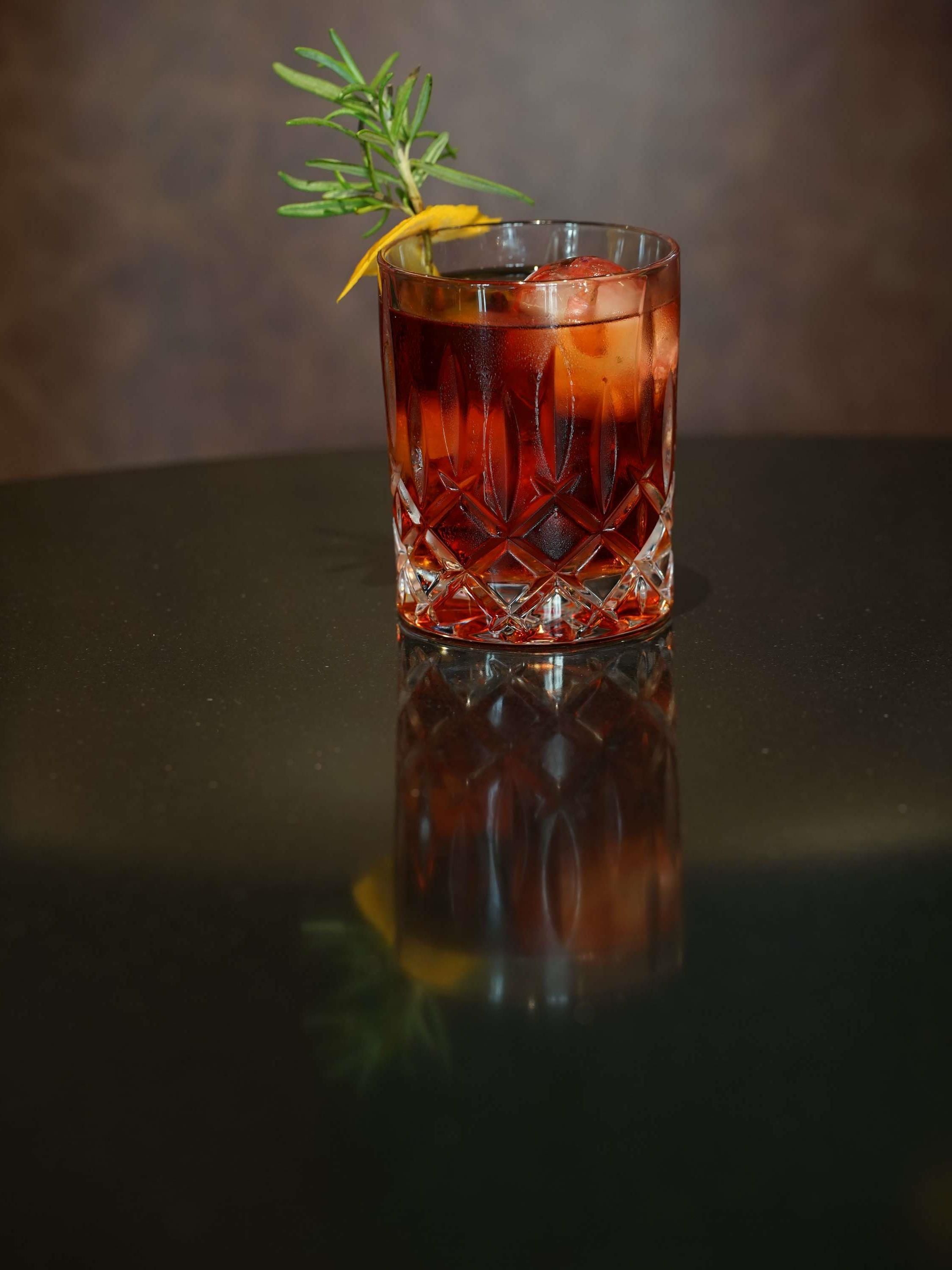 A glass of amber-colored beverage with ice cubes and a sprig of rosemary on a reflective surface.