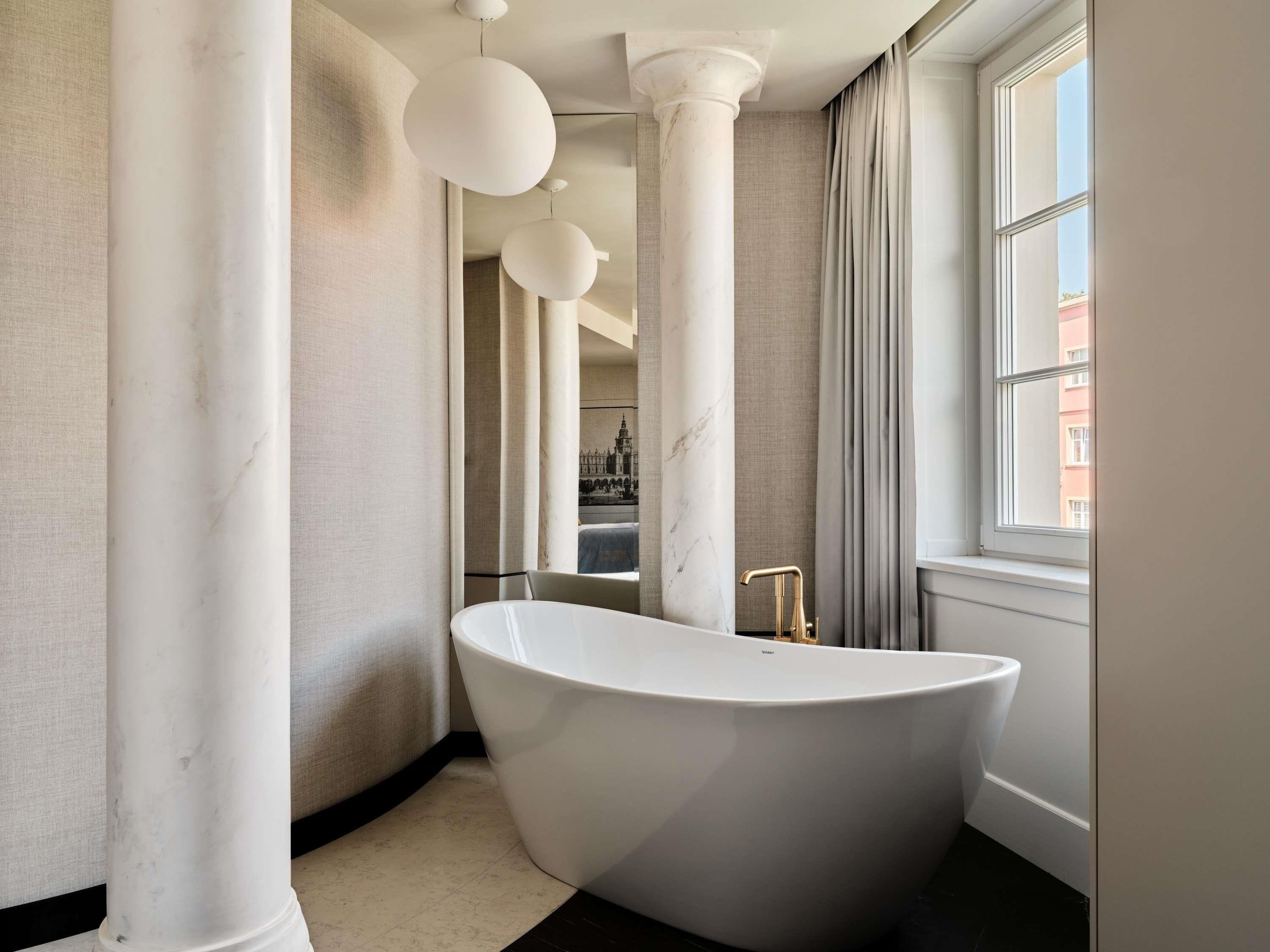 A modern bathroom with a freestanding bathtub, marble columns, and a large window with curtains.