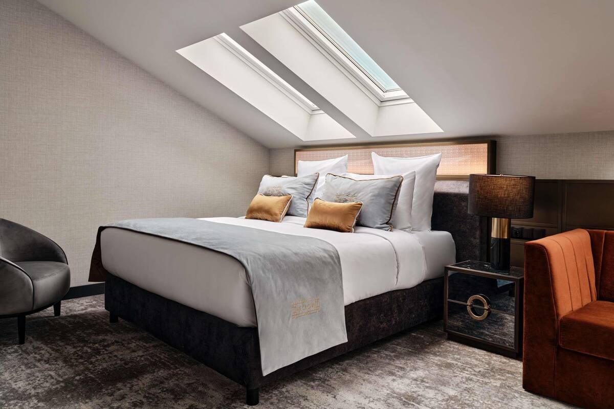 Ein modernes Schlafzimmer mit Dachfenster, einem Bett mit vielen Kissen, einem Nachttisch, einer Lampe und einem Sessel.