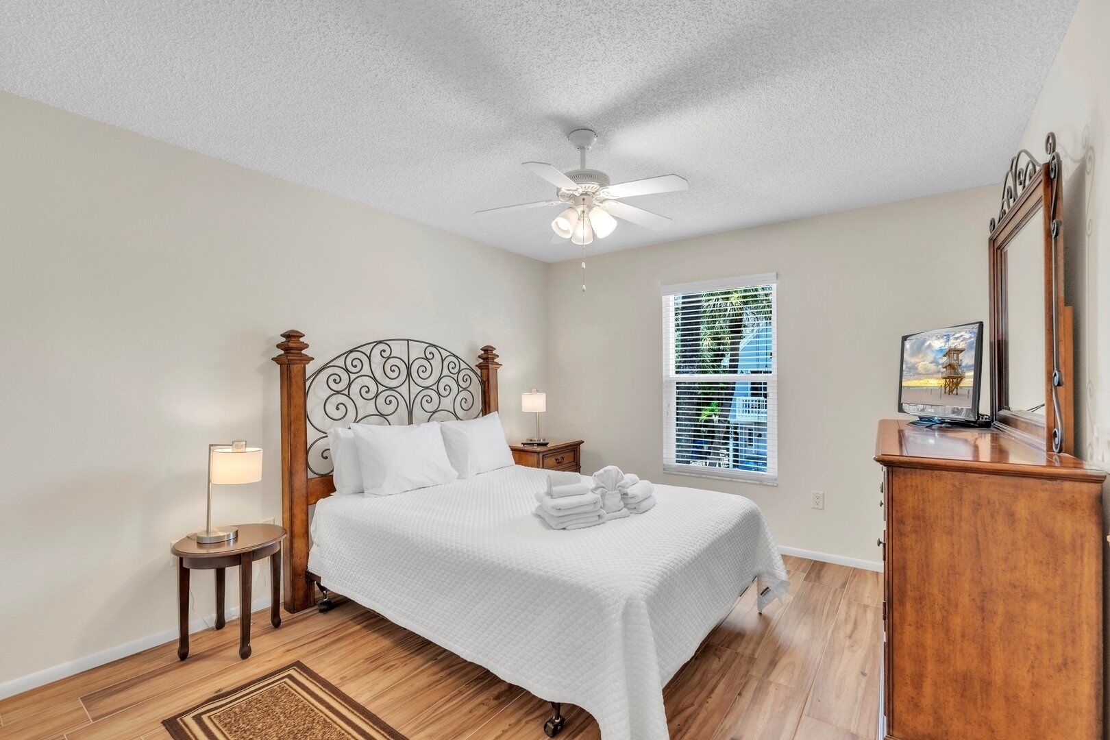 A bedroom with a bed, a ceiling fan, a nightstand, a lamp, a window with blinds, and a wooden dresser with a TV.