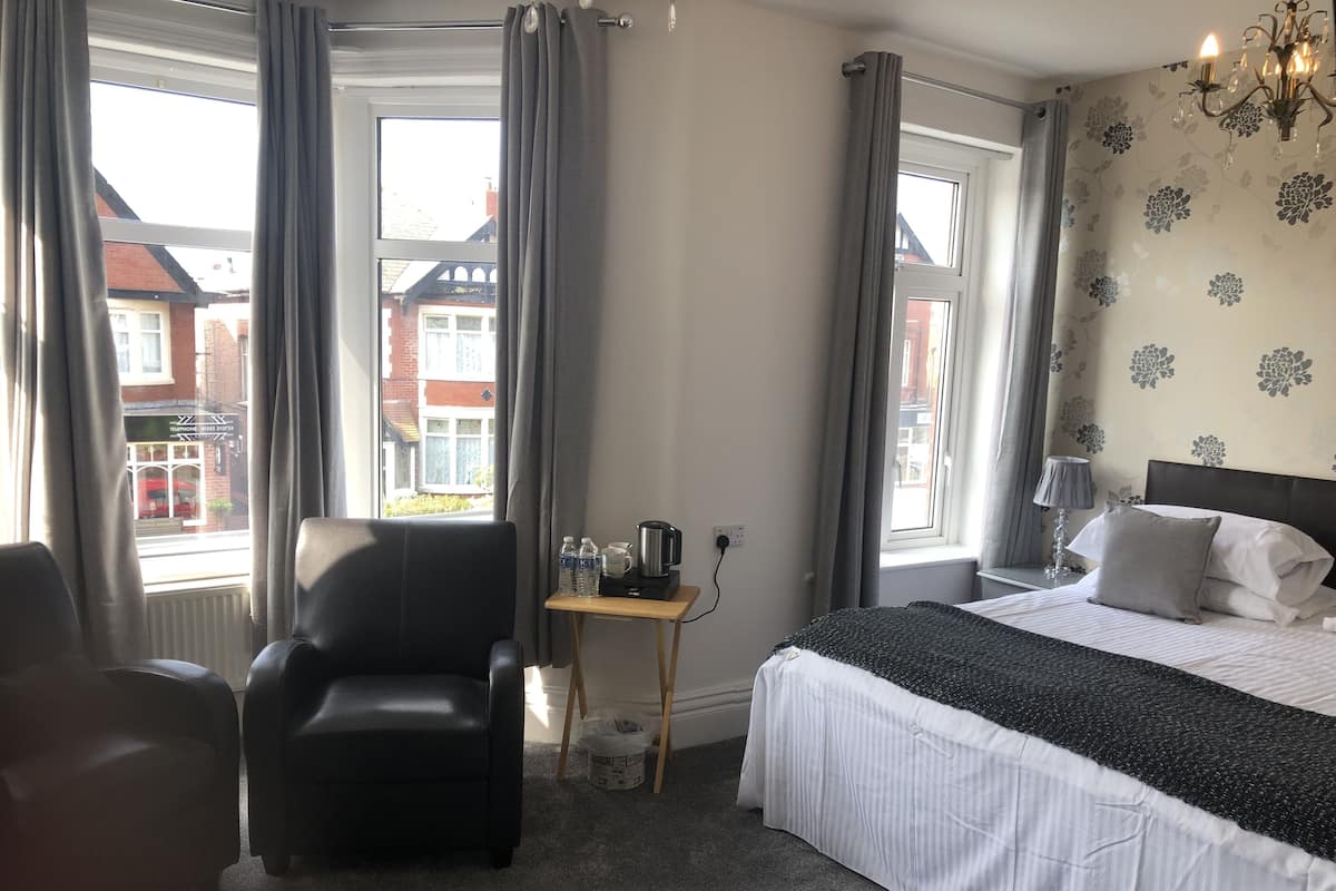 A hotel room with a bed, a chair, a small table with a kettle and water bottles, and a view of buildings outside.