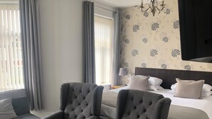 Una habitación de hotel con una cama, dos sillones, una mesita de noche, una lámpara y un televisor de pared.