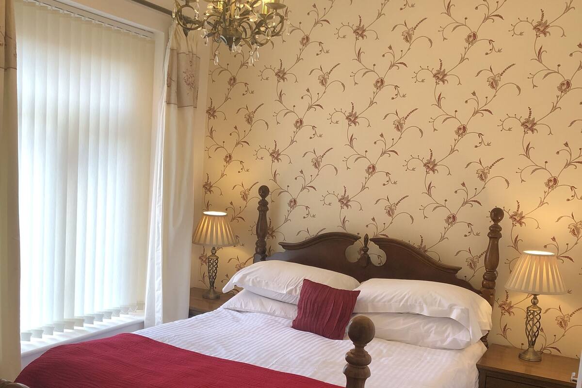 A neatly made bed with a wooden headboard, white and red bedding, two bedside lamps, and a floral wallpaper pattern.