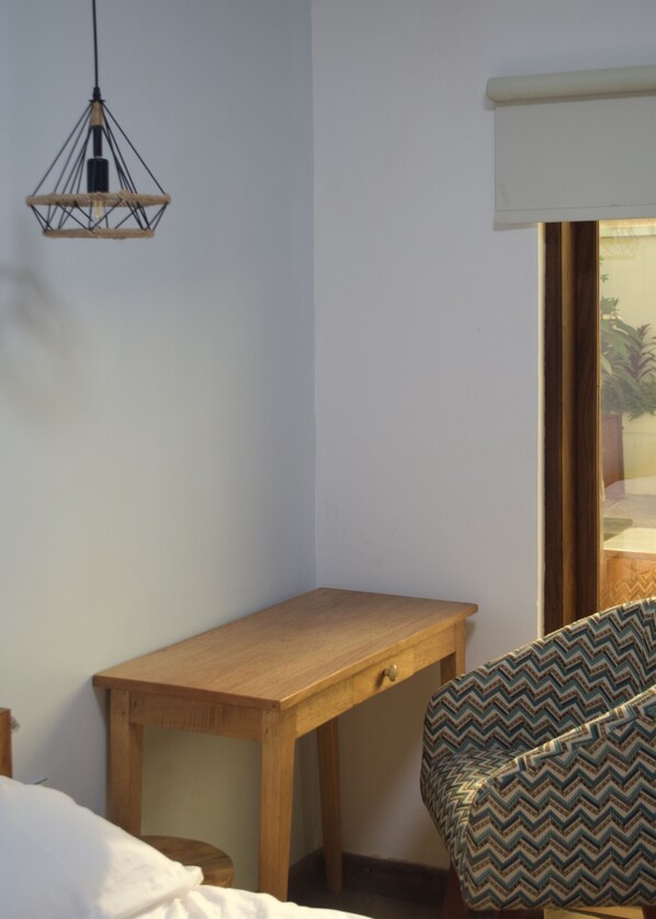 A small room with a wooden desk, a chair with a patterned cushion, a hanging light fixture, and a window with a view of greenery.