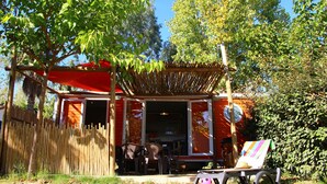 Un área de comedor al aire libre de estilo rústico, con una cerca de madera, un edificio rojo con puerta abierta y una pérgola de madera.