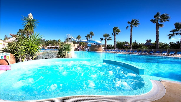 Una amplia piscina exterior con jacuzzi, rodeada de palmeras y sillones de descanso.
