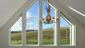 Une pièce avec un plafond en pente et un lustre. Trois grandes fenêtres offrent une vue sur un champ verdoyant.