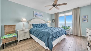 Un dormitorio con cama, mesitas de noche, una silla, ventilador de techo y vista al océano.