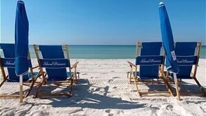 Cuatro sillas de playa con fundas azules y estructura de madera, dispuestas en una playa arenosa con vista despejada al océano.