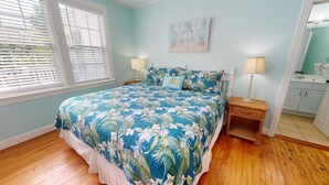 Une chambre à coucher avec une couvre-lit à motifs floraux, une table de chevet avec une lampe, un tableau accroché au mur et une salle de bain visible à travers une porte ouverte.
