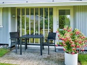 Een overdekt terras met een tafel en twee stoelen, een potplant met rode bloemen en uitzicht op groen door de glazen deuren.